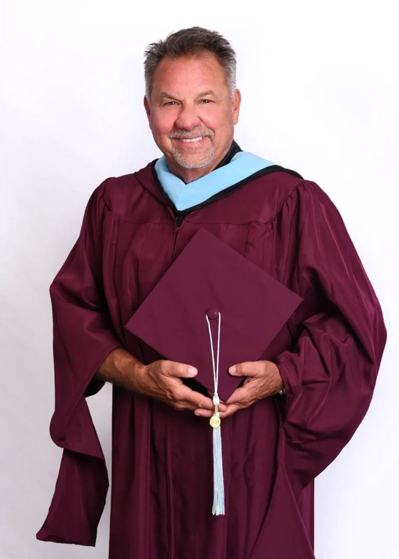 A man in graduation gown holding his cap and diploma.